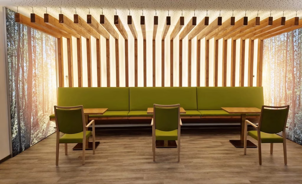 Grüne Sitzbank und Stühle in einer Sitzecke mit Holzlamellen und Waldmotiven an den Wänden.