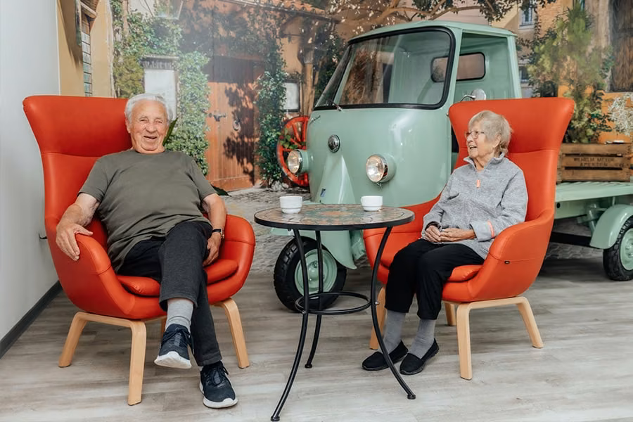 Ein Mann und eine Frau sitzen mit Kaffeetassen in roten Sesseln vor einem grünen Dreiradfahrzeug.